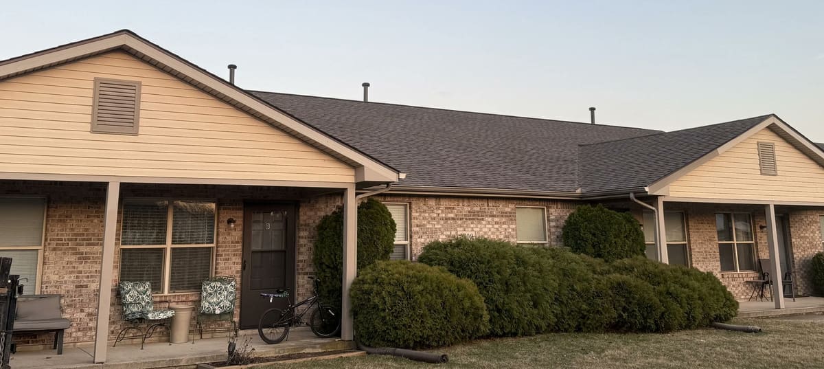 Tan brick ranch with mature shrubs and a completed brown shingle roof