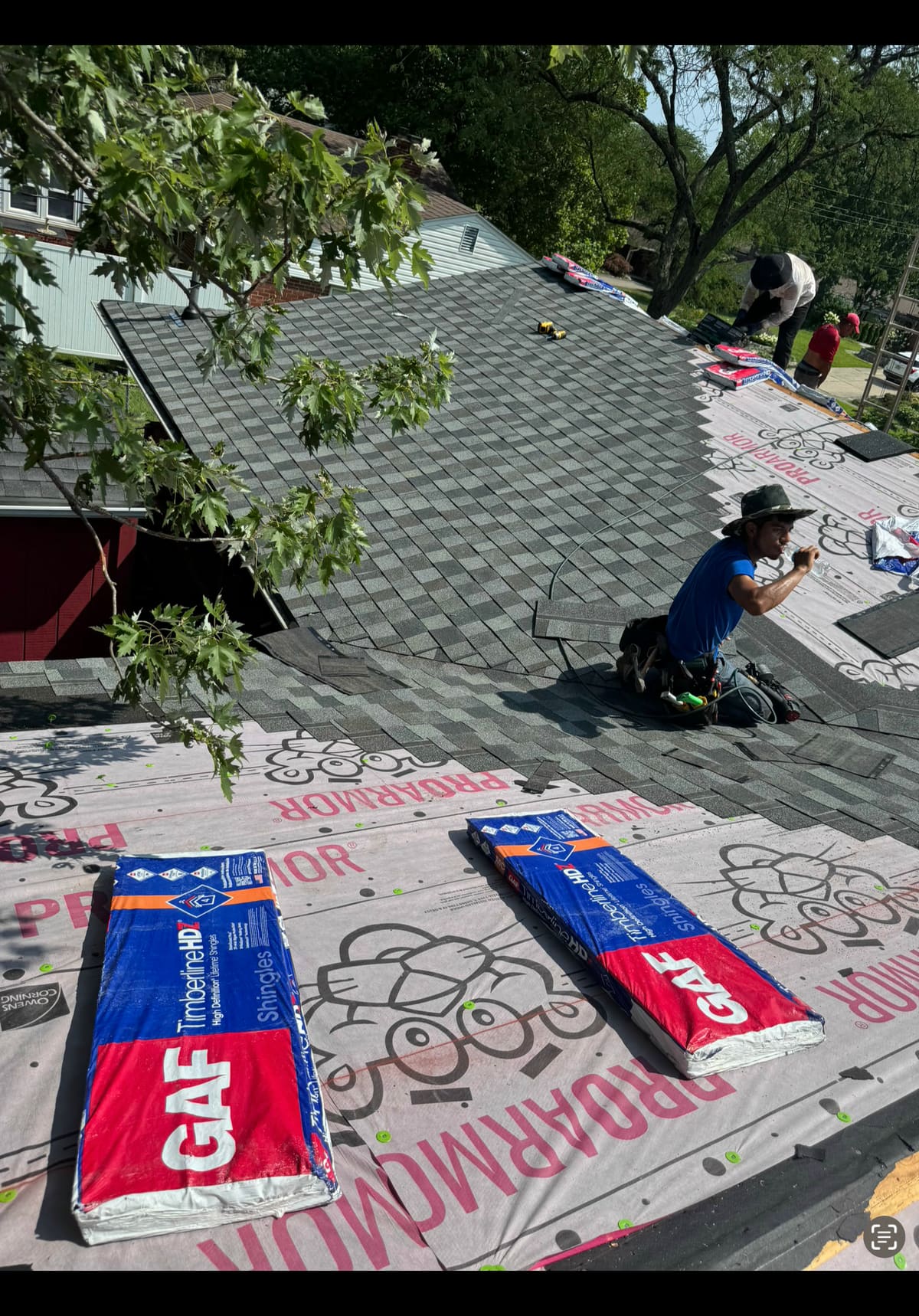 Roofer kneeling on synthetic underlayment with shingle bundles staged across the deck