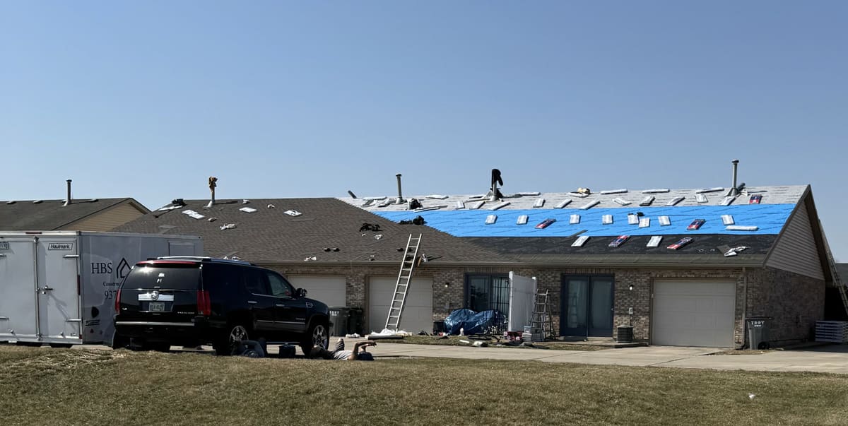 Long roof stripped back mid tear-off with a blue tarp, ladder and debris containment below