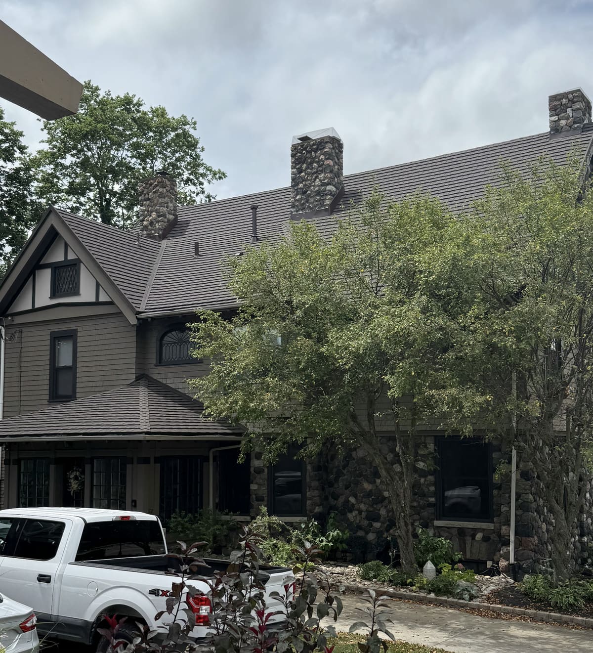 Dark two-story older home with twin stone chimneys, seen through a mature tree